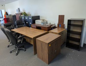 Another view of used office chairs, desks, shelves, filing cabinets and a photocopier wait in a white-walled office area, to be taken away by Furniture Bank